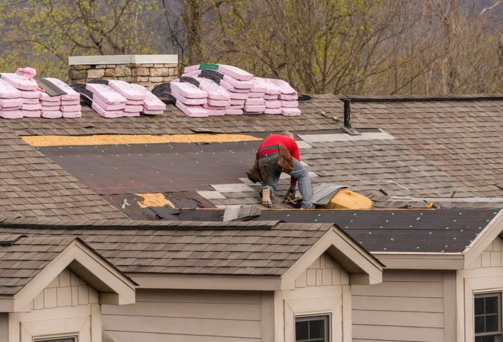 A roofer repairing a shingle roof, installing new underlayment and shingles while insulation bundles are stacked near the chimney.