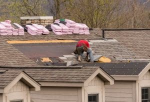 A roofer repairing a shingle roof, installing new underlayment and shingles while insulation bundles are stacked near the chimney.