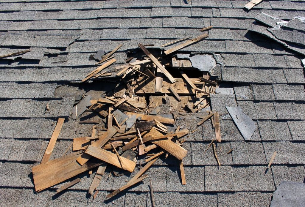 A damaged shingle roof with a large hole exposing the underlying wooden structure and scattered broken roofing materials.